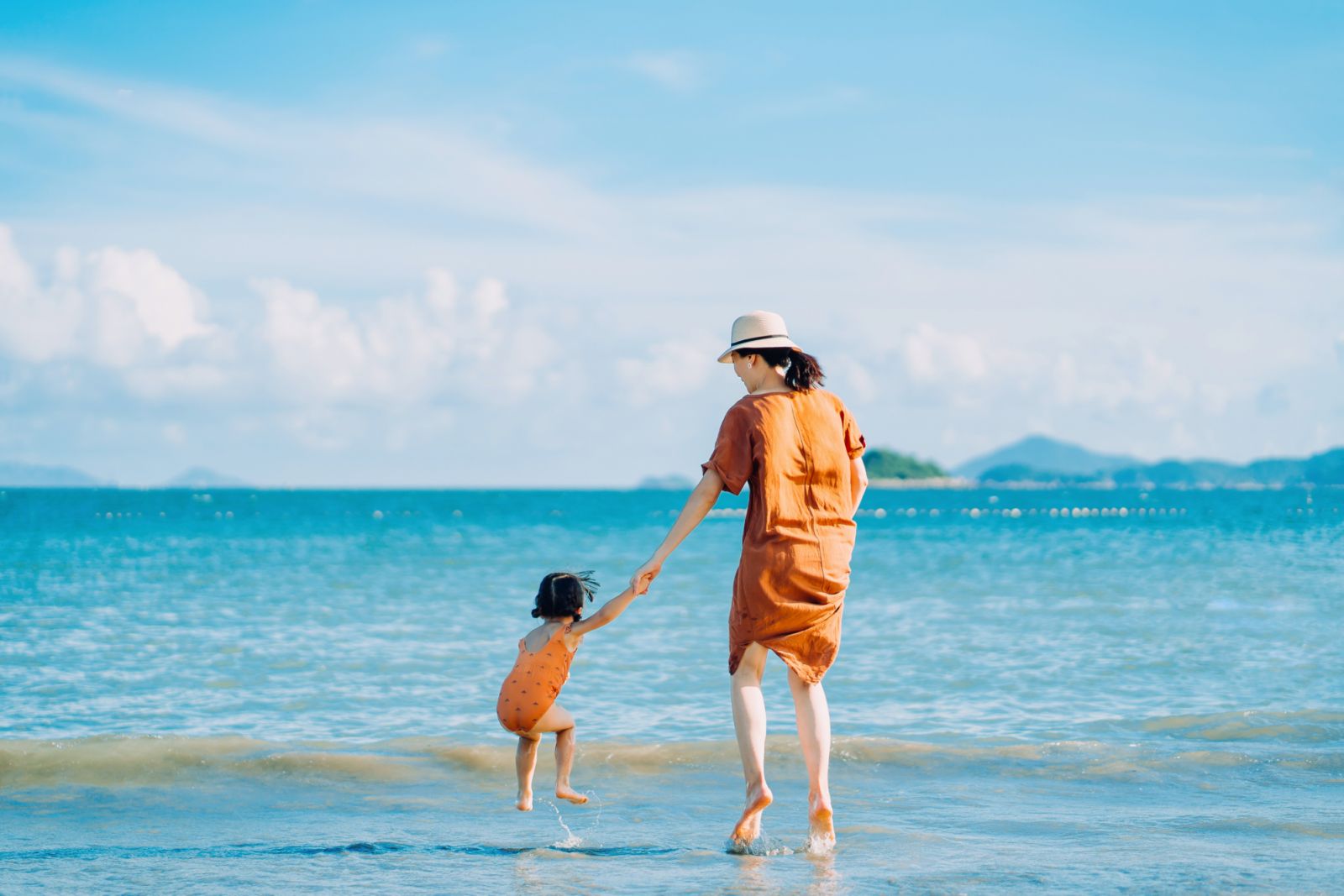 Rear view of joyful young Asian mother and little daughter holding hands while jumping on the beach against blue sky on a sunny Summer day